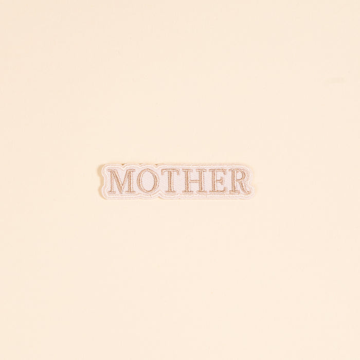 The Darling Effect’s Mother Mix & Match Iron-On Patch features “MOTHER” in tan serif on white with a scalloped edge. Perfect for customizing your items, it’s shown against a cream background.