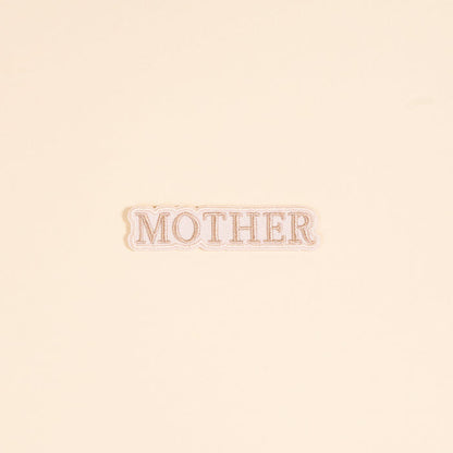 The Darling Effect’s Mother Mix & Match Iron-On Patch features “MOTHER” in tan serif on white with a scalloped edge. Perfect for customizing your items, it’s shown against a cream background.