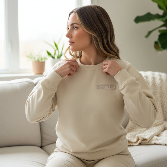 A woman sits on a light sofa in a beige sweatshirt reading "MOTHER," ready to personalize items with The Darling Effect's Mother Mix & Match Iron-On Patch. She looks to the side; plants and a knitted blanket are seen as sunlight streams in.