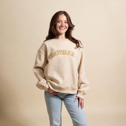 A smiling young woman showcases The Darling Effect’s Mother Signature Crew sweatshirt in beige with gold "MOTHER" lettering and light blue jeans against a cream background, highlighting this soft essential piece.