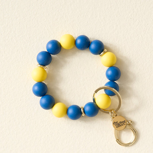 The Darling Effect’s Navy Yellow Game Day Hands-Free Wristlet features alternating blue and yellow beads, a gold ring, and a small charm engraved with “The Darling Effect,” arranged on a light cream background.