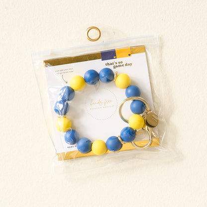 The Darling Effect Navy Yellow Game Day Hands-Free Wristlet features large blue and yellow beads, a gold keyring, and comes in a clear bag with a white “that’s so game day” card with gold accents—perfect for school pride.
