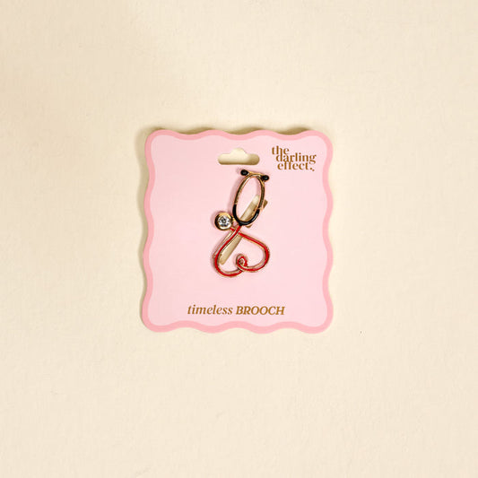 The Nurse Love Timeless Brooch by The Darling Effect, a gold and pink vintage-style heart design with a small gem, is shown on a scalloped pink card labeled “the darling effect. timeless BROOCH” against a light background.