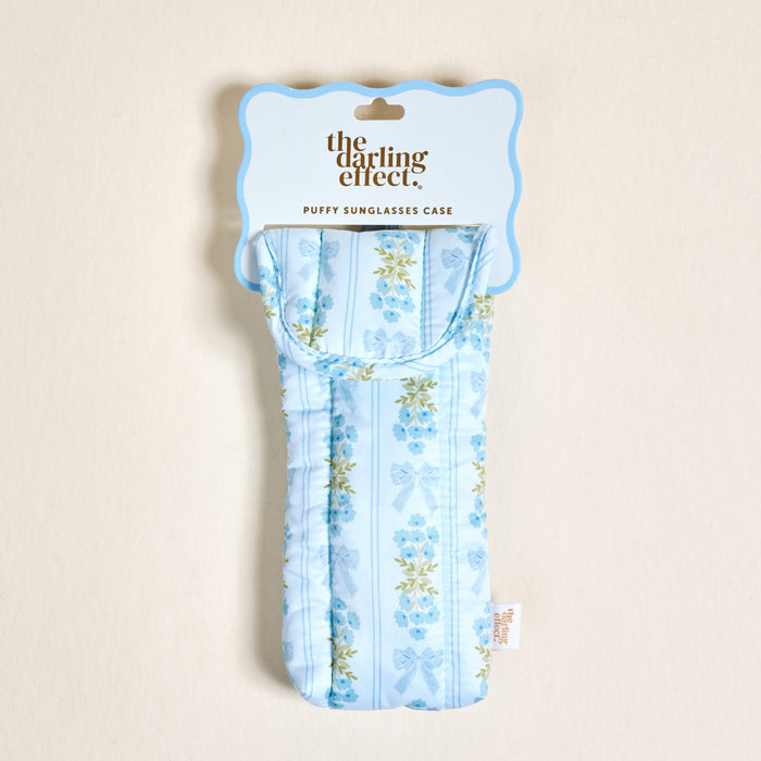 The Oh So Fancy Blue Puffy Sunglasses Case by The Darling Effect features a floral and ribbon pattern, magnetic flap closure, and comes packaged with a scalloped-edge label reading “the darling effect. PUFFY SUNGLASSES CASE.”.