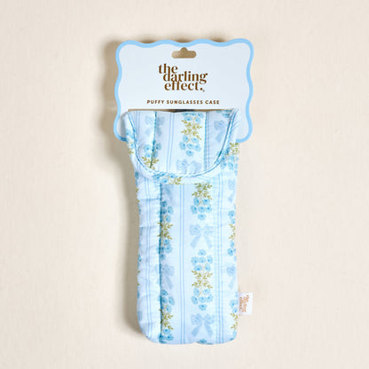 The Oh So Fancy Blue Puffy Sunglasses Case by The Darling Effect features a floral and ribbon pattern, magnetic flap closure, and comes packaged with a scalloped-edge label reading “the darling effect. PUFFY SUNGLASSES CASE.”.