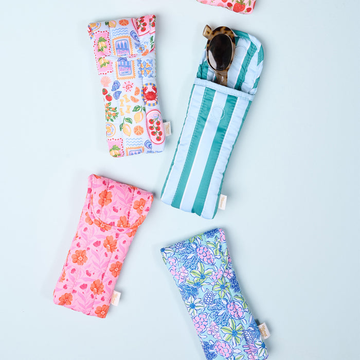 Five Oh So Fancy Blue Puffy Sunglasses Cases by The Darling Effect, featuring floral and geometric patterns with magnetic flap closures, are arranged on a light blue background. One case is open, showing sunglasses inside.