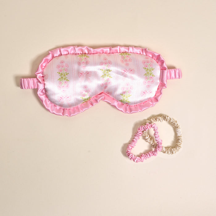 The Darling Effect’s Oh So Fancy Pink Beauty Sleep Mask with floral print and ruffled trim is paired with two matching satin scrunchies, creating a cozy sleep set on a light beige background.