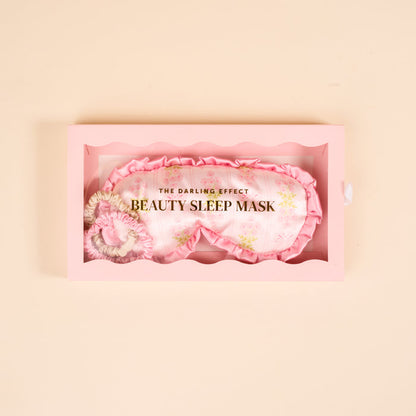 A wavy-edged pink box holds The Darling Effect’s Oh So Fancy Pink Beauty Sleep Mask—a light pink satin mask with a floral pattern and gold text: THE DARLING EFFECT and BEAUTY SLEEP MASK.