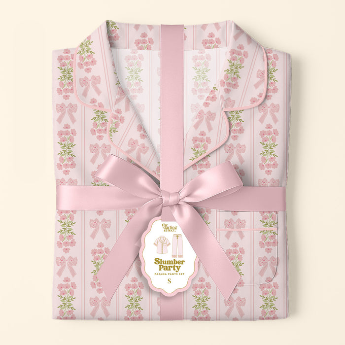 Folded Oh So Fancy Pink Slumber Party Pajama Pants Set by The Darling Effect, featuring a floral and bow pattern in ultra-soft fabric, tied with a pink satin ribbon and gift tag labeled "Slumber Party Pajama Set, S," on a light background.