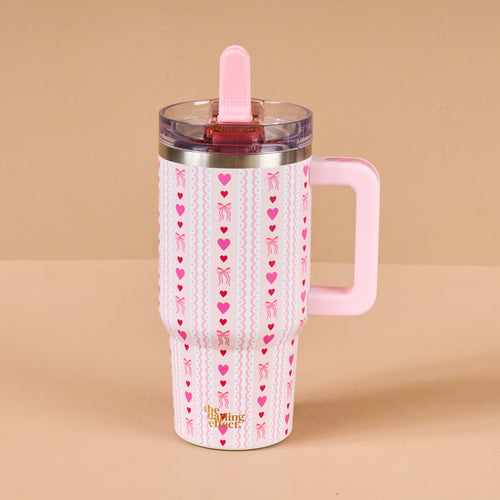The Darling Effect's Oh So Lovely 20 oz Lifestyle Flip Straw Tumbler features pink stainless steel, a handle, hearts, bows, vertical stripes, and a transparent lid with a pink slide. This Valentine’s mug is shown on a beige background.