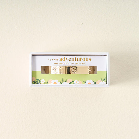A box featuring The Darling Effect's Original Trait Cuff Bracelet, engraved with inspiring designs, displayed on a floral background. Packaging message: "You are adventurous. Seek the road less traveled.