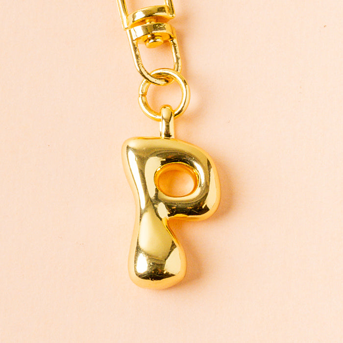 The-darling-effect P Bubble Letter Keychain Bag Charm features a shiny gold letter "P" pendant on a gold keychain, displayed on a peach background.