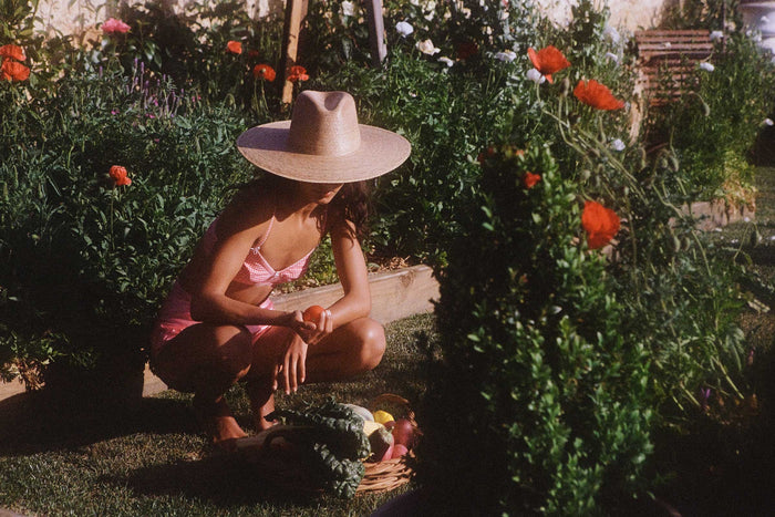 A woman wearing a Palma Wide Fedora in a garden, crouching with a basket of vegetables.