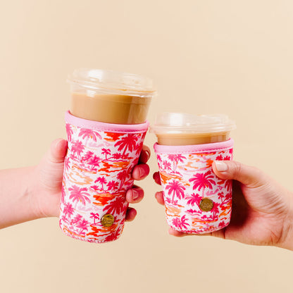 Two hands hold iced coffee cups with Paradise Palms Sip Savvy Drink Sleeves by the-darling-effect, featuring pink and orange tropical palm patterns and small gold tags, against a beige background. Reusable and stylish for your drinks.