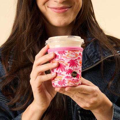 A woman in a denim jacket smiles gently while holding a plastic cup with the-darling-effect’s Paradise Palms Sip Savvy Drink Sleeve, featuring pink and orange palm tree patterns.