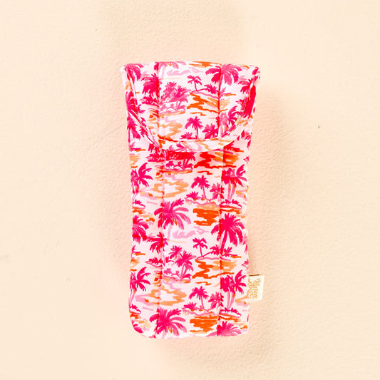 The Paradise Palms Sunglasses Case by the-darling-effect is a soft, quilted case featuring a pink, orange, and red palm tree pattern on a light background, shown against a pale peach surface.