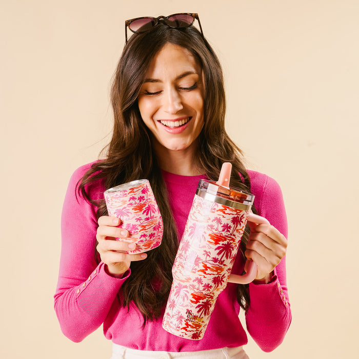 A woman in a pink sweater smiles, holding two Paradise Palms Wine Tumblers by The Darling Effect with pink and orange palm tree designs. Sunglasses rest on her head. Each tumbler features triple-wall insulation for ideal drink temperature.