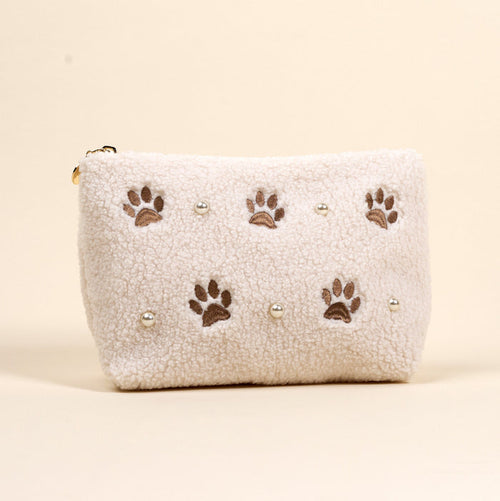 The Darling Effect’s Paws & Pearls Teddy Pouch is a fluffy white sherpa accessory featuring embroidered brown paw prints and pearl accents.