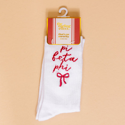 A pair of Pi Beta Phi Embroidered Crew Socks by The Darling Effect, featuring white fabric, pink bow, and sorority name on a peach background—perfect for showing off your Pi Beta Phi spirit.
