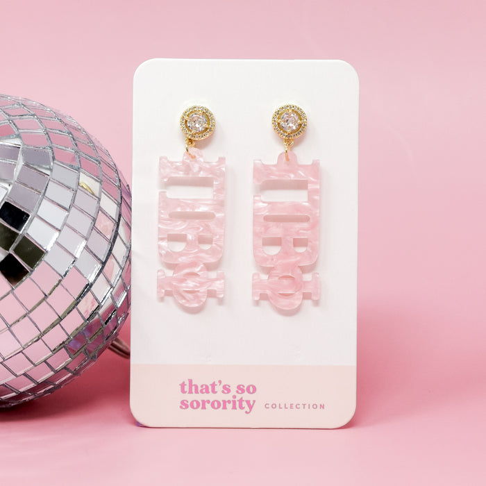 A pair of Pi Beta Phi Letter Pink Acrylic Earrings by The Darling Effect hang on a display card, set against a pink background with a disco ball—perfect for sisterhood events or adding to a bid-day basket.