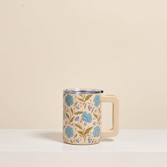 The Darling Effect's Poppin' Petals Aqua 14 oz Insulated Mug, featuring blue flowers and green leaves, with a large handle, sits on a white surface against a plain beige background.