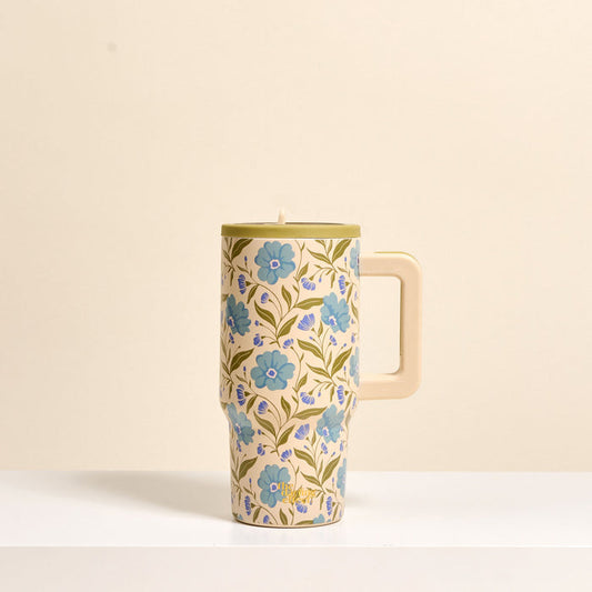 The Darling Effect’s Poppin' Petals Aqua 20 oz Lifestyle Flip Straw Tumbler, with a large handle and a floral design of blue and purple flowers with green leaves on cream, sits on a white surface against a beige backdrop.