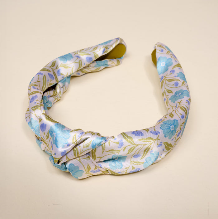The Darling Effect’s Poppin' Petals Aqua Classic Twist Headband features light, silky fabric with blue flowers and green leaves on a pale background—an elegant, stylish accessory shown on a neutral surface.