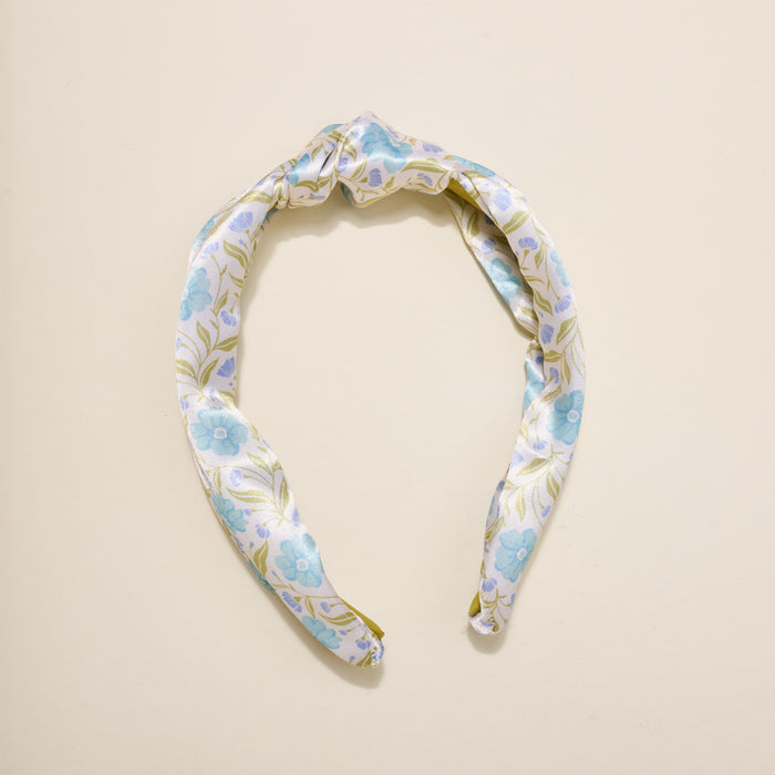 The Darling Effect’s Poppin' Petals Aqua Classic Twist Headband, featuring a blue floral pattern and elegant top knot, rests on a cream background—making it a charming hair accessory.