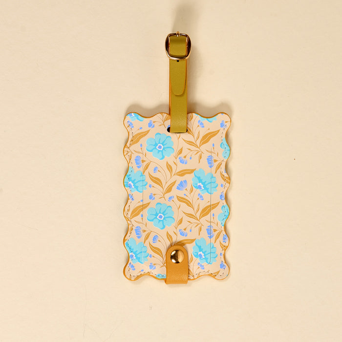 The Darling Effect's Poppin' Petals Aqua Jetsetter Luggage Tag features a rectangular, wavy-edged design with a light beige background, blue flowers, gold leaves, privacy flap, and a green strap with a gold buckle.