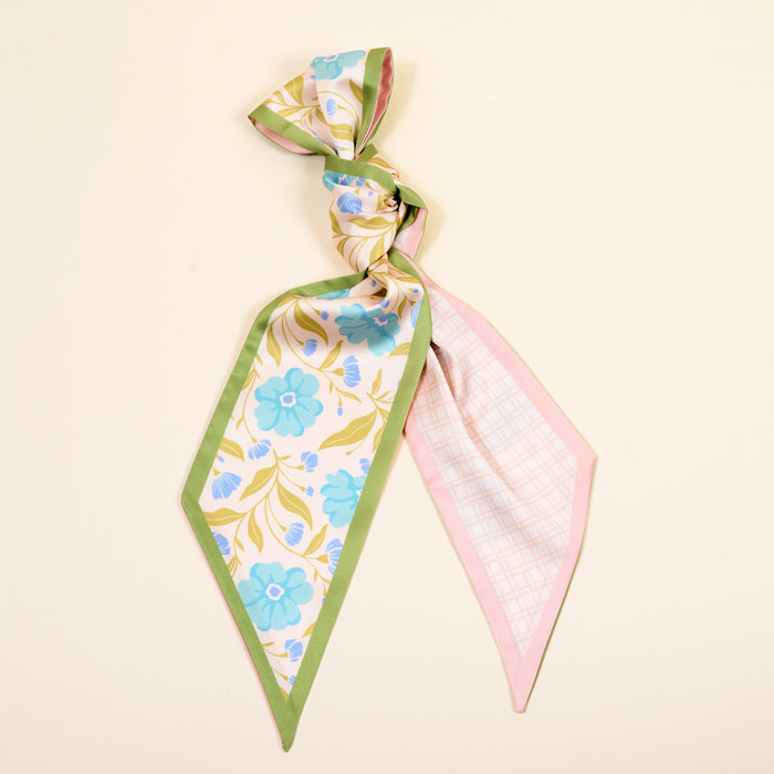 The Darling Effect’s Poppin' Petals Aqua Twirl & Tie Scarf Accessory features a green border, blue flowers, yellow leaves on one side, and pink plaid on the other—add boutique charm and versatile style by tying it in a loose knot.