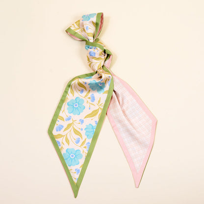 The Darling Effect’s Poppin' Petals Aqua Twirl & Tie Scarf Accessory features a green border, blue flowers, yellow leaves on one side, and pink plaid on the other—add boutique charm and versatile style by tying it in a loose knot.