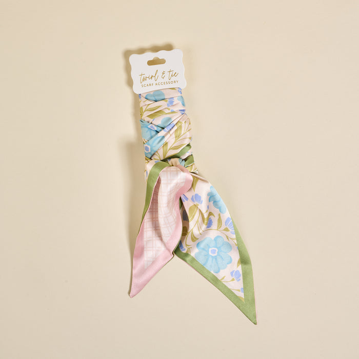 The Poppin' Petals Aqua Twirl & Tie Scarf Accessory by The Darling Effect, featuring floral patterns in pink, blue, and green, is showcased on a beige background with a white “Touched & Free Scarf Accessory” tag—perfect for versatile styling.