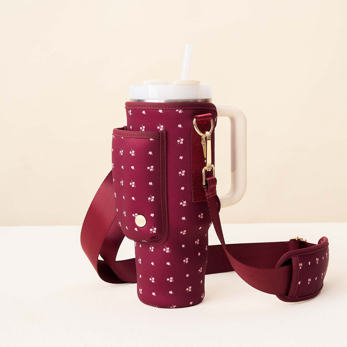 The Darling Effect’s Pretty Posies Plum Tumbler Sling, with a maroon and white floral pattern, holds a large mug with handle and straw. It features an adjustable strap, pocket, and gold buckle, set on a white surface with a beige background.