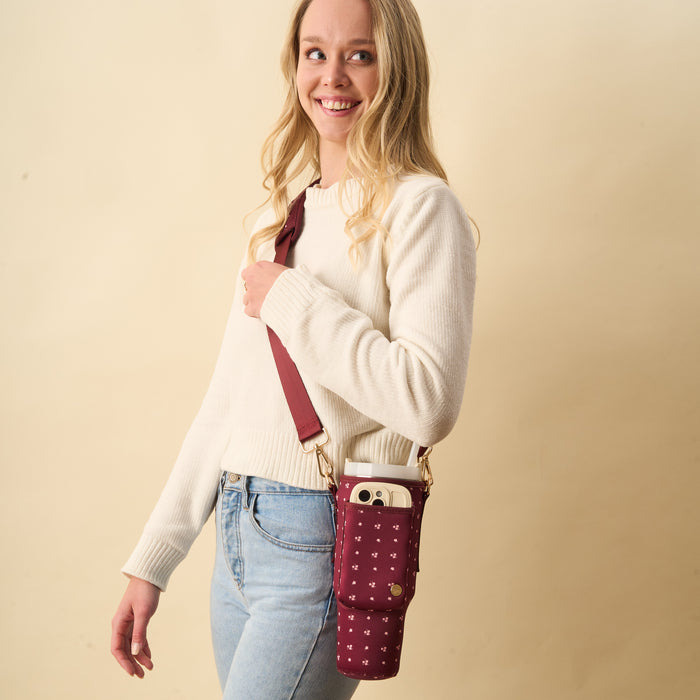 A smiling woman with long blonde hair wears a white sweater and light blue jeans, carrying The Darling Effect's Pretty Posies Plum Tumbler Sling with an adjustable strap. Its front pocket holds a phone and tumbler as she stands by a beige background.