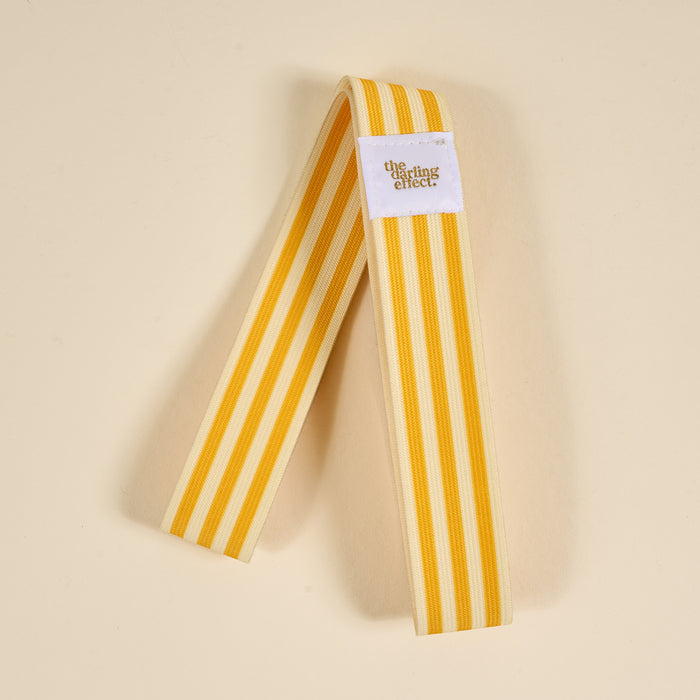 The Darling Effect Put Towel Band-Stripes Yellow Stay features yellow and cream stripes, a white tag with the logo, and works as a stylish, no-slip towel holder for your beach chair on sunny days.