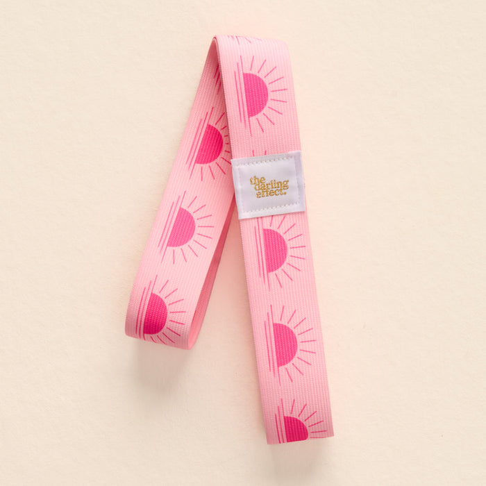 The-darling-effect Put Towel Band-Sunshine Pink Stay is a pink fabric resistance band with a half-sun pattern and gold-accented label. It also secures no-slip towels stylishly to any beach chair.