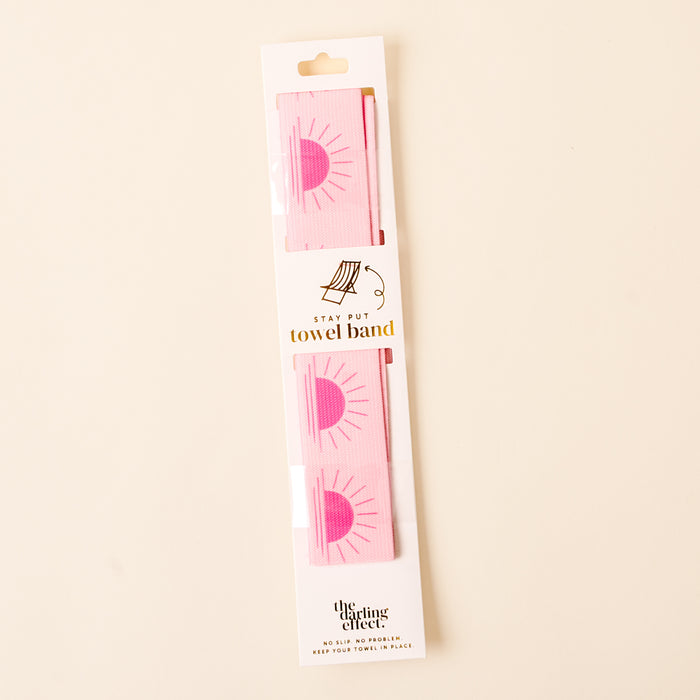 A packaged Put Towel Band-Sunshine Pink Stay by the-darling-effect, designed as a no-slip towel holder with a pink sun design; white packaging features gold and black text plus the brand name at the bottom. Perfect for beach chairs!.