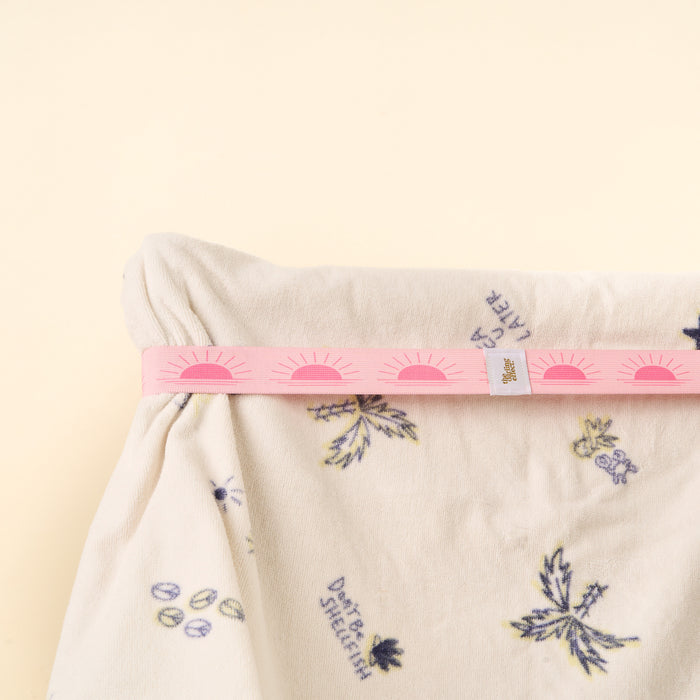 The-darling-effect's Put Towel Band-Sunshine Pink Stay secures a cream fabric with purple and yellow flowers. The pink straps have repeating sun motifs, all set against a plain beige background.