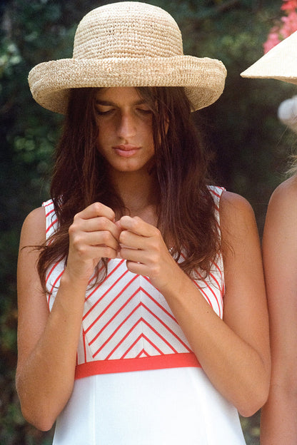 Model wearing a Raffia Cruiser straw boater hat, with a white dress and red stripes, outdoors.