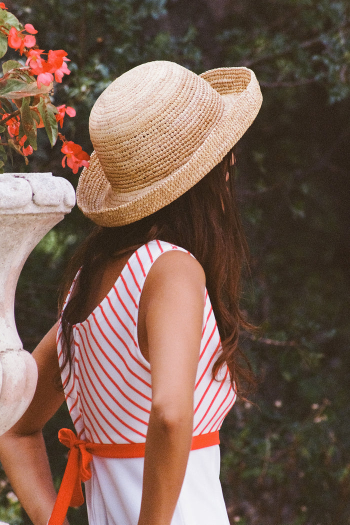A woman wearing a Raffia Cruiser straw boater hat, with a natural woven texture, standing near flowers.