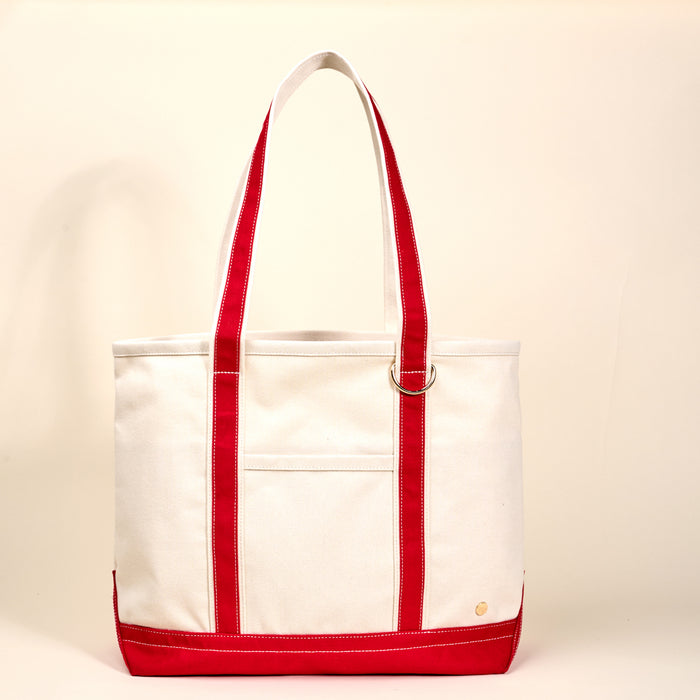 The Red The Signature Canvas Tote Bag by The Darling Effect features durable red handles, trim, and base, an exterior pocket, and a gold emblem on white canvas—ideal for everyday use.
