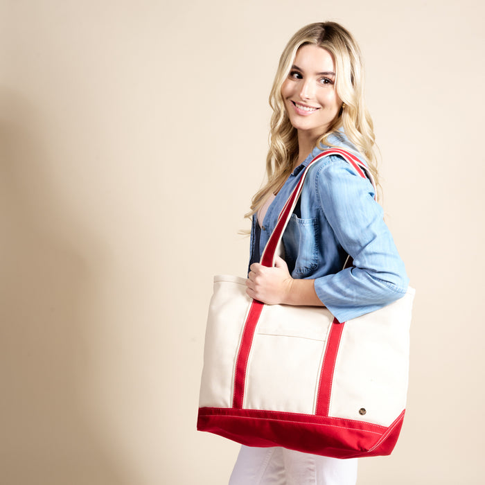 A woman with long blonde hair, in a light blue shirt and white pants, smiles while holding The Darling Effect’s Red The Signature Canvas Tote Bag over her shoulder against a beige background.