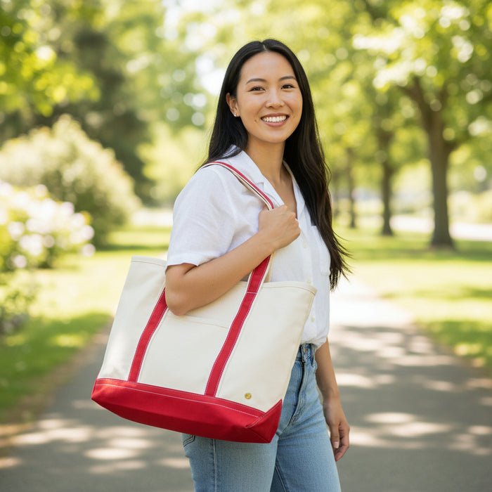 A woman with long dark hair, in a white shirt and jeans, smiles outdoors on a sunny day. She carries The Darling Effect’s Red The Signature Canvas Tote Bag with red straps—an everyday essential. Trees and greenery fill the background.