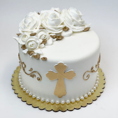 A decorated cake with white buttercream icing, white buttercream roses, gold fondant cross, and gold scroll patterns on a white background.