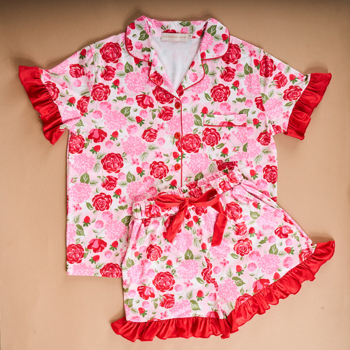 The Darling Effect's Rose Garden Slumber Party Pajama Shorts Set features ultra-soft fabric with red and pink rose prints, green leaves, a relaxed fit, short-sleeved top, shorts with red ruffled trim and ribbon bow—perfect for slumber parties.