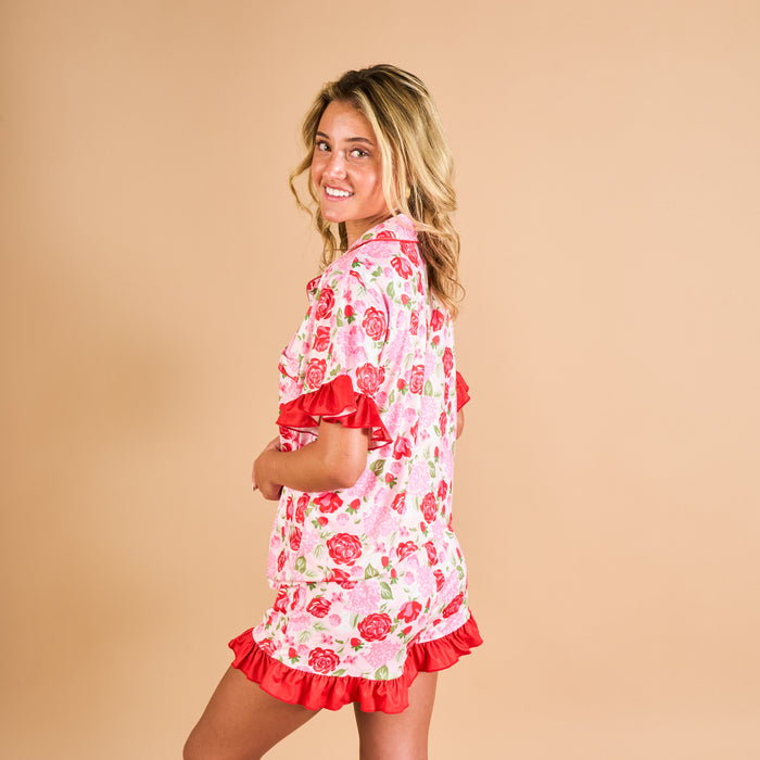 A young woman smiles over her shoulder, wearing The Darling Effect’s Rose Garden Slumber Party Pajama Shorts Set—a pink set with red rose print, red ruffles, and a relaxed fit.