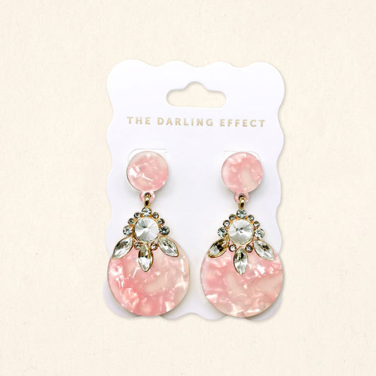 The Darling Effect Rosé Radiance Show Stopper Earrings feature pink marble acrylic teardrops, crystal rhinestones, gold-tone accents, and round studs. Lightweight and eye-catching, they come displayed on a branded jewelry card.