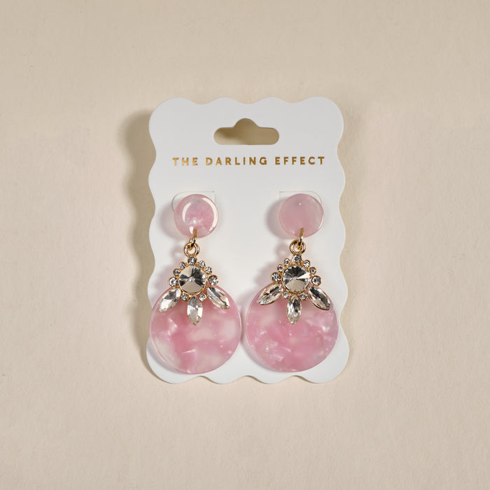 The Rosé Radiance Show Stopper Earrings by The Darling Effect are pink, lightweight statement earrings with rhinestone accents, displayed on a white card with gold branding against a neutral beige backdrop.