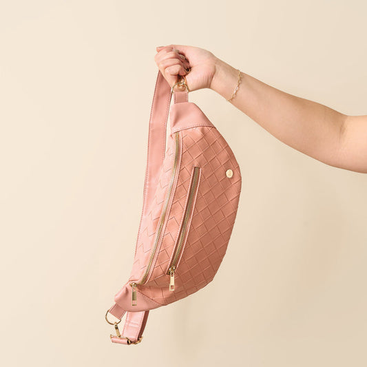 A hand holds The Darling Effect’s Rose Trendy Luxe Belt Bag, crafted from blush pink vegan leather with gold zippers, against a cream background.