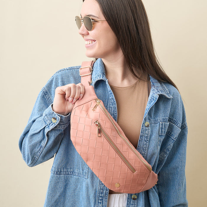 A young woman with long dark hair, sunglasses, and a denim jacket smiles while holding The Darling Effect’s Rose Trendy Luxe Belt Bag in woven pink vegan leather.